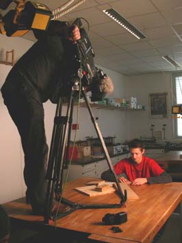 Foto der Dreharbeiten. Auf der Werkbank steht ein Kameramann mit Stativ und Kamera. Ich sitze am Tisch mit Holzproben